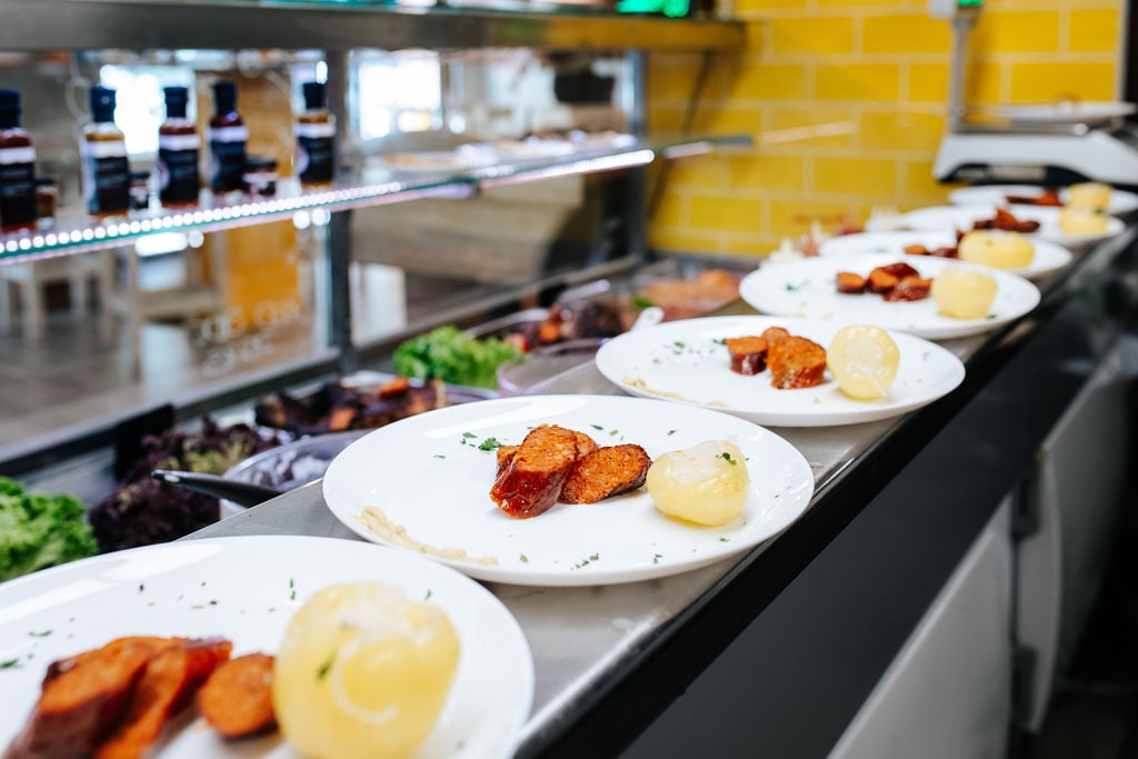 Slices of traditional Hungarian smoked sausage and a pickled apple pepper served on white plates lined up on a restaurant counter.